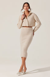 A woman is wearing a beige, cropped, zip-up fuzzy jacket with a high collar and a matching high-waisted, fitted pencil skirt that falls below the knees. She is also wearing a pair of beige pointed-toe heels with a bow detail on the front.